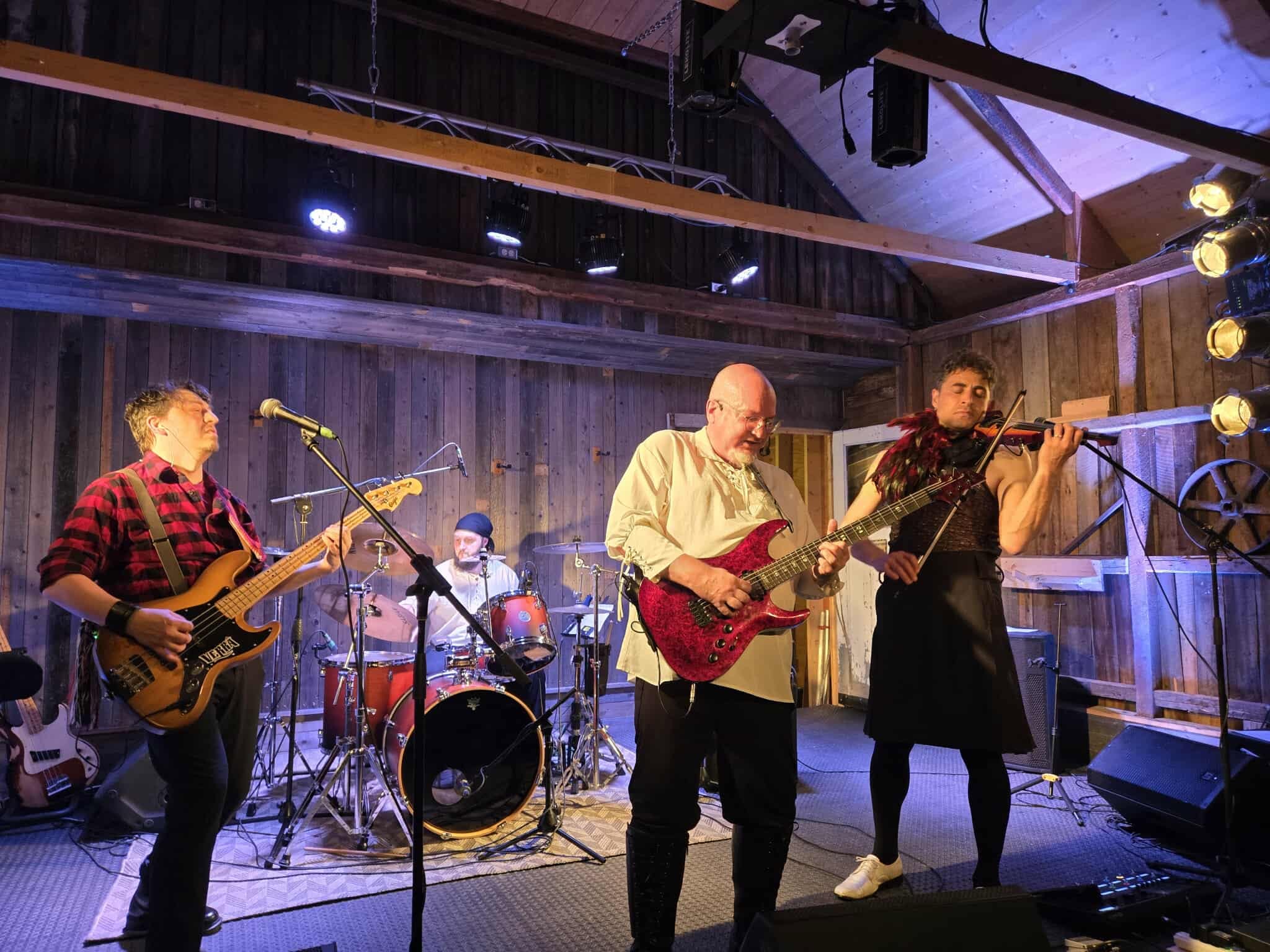 MôdiVerrâ performing together in a wood-paneled venue: bass, drums, electric guitar and violin under stage lights.